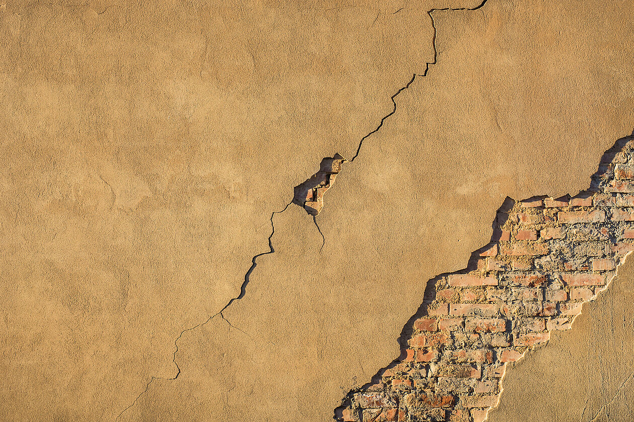 Nahaufnahme einer verputzten Außenwand mit langem Riss und abgeplatztem Putz, darunter freiliegendes Mauerwerk.