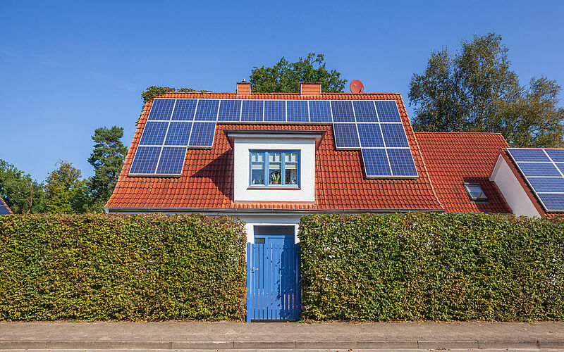 Ein modernes Einfamilienhaus mit rotem Ziegeldach und großflächigen Solarpaneelen steht hinter einer gepflegten Hecke unter klarem, blauem Himmel.