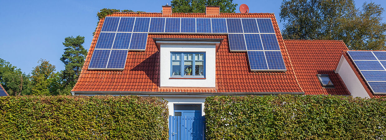 Ein modernes Einfamilienhaus mit rotem Ziegeldach und großflächigen Solarpaneelen steht hinter einer gepflegten Hecke unter klarem, blauem Himmel.