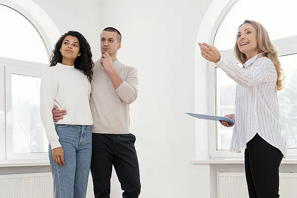 Ein junges Paar steht in einer leeren Wohnung mit großen Rundbogenfenstern, während eine Immobilienmaklerin ihnen etwas erklärt und in den Raum zeigt. Die Szene wirkt wie eine Wohnungsbesichtigung.