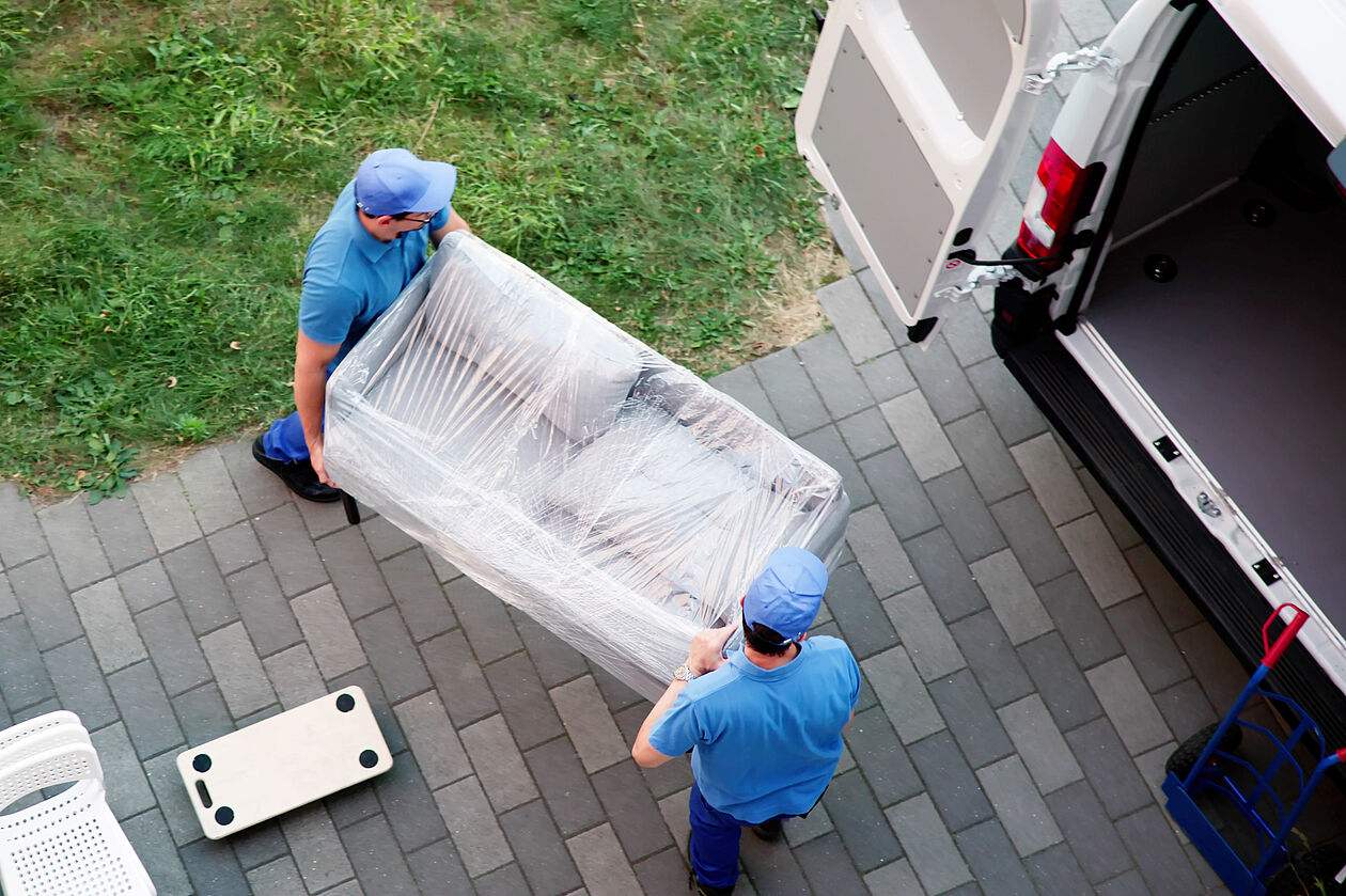 Zwei Umzugshelfer tragen eine verpackte Couch in einen Lieferwagen.