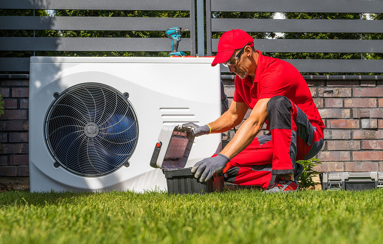 Installateur in roter Arbeitskleidung arbeitet im Garten an einer Luft-Wasser-Wärmepumpe.