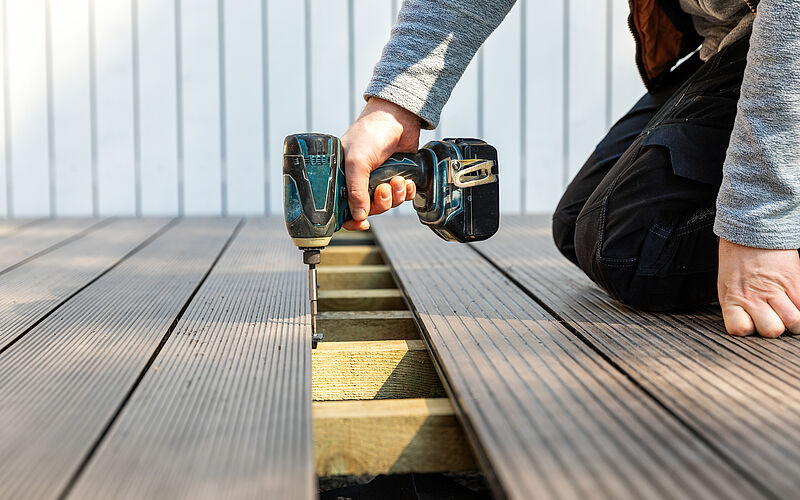 Eine Person kniet auf einer Terrasse und setzt den Bohrer zwischen zwei Terrassendielen an.