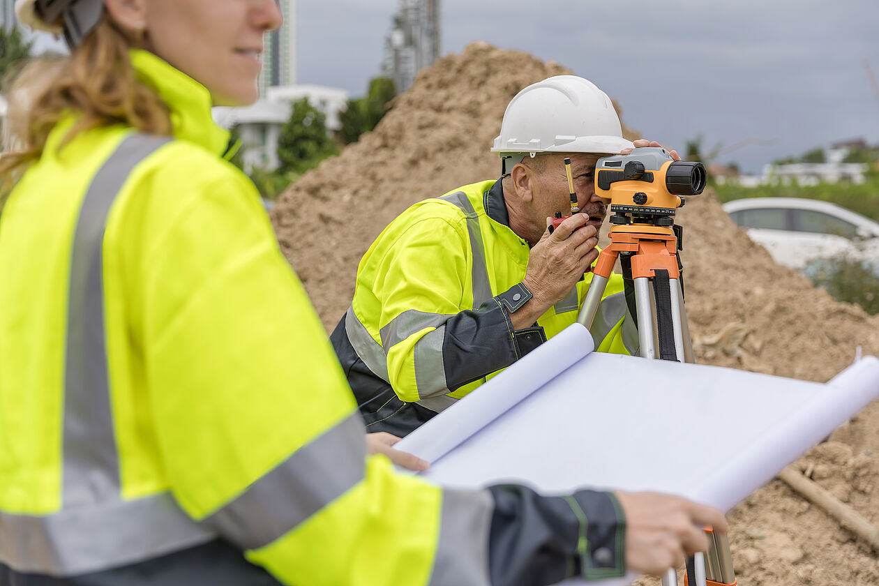 Techniker bei der Grundstücksvermessung mit Nivelliergerät auf Baustelle.