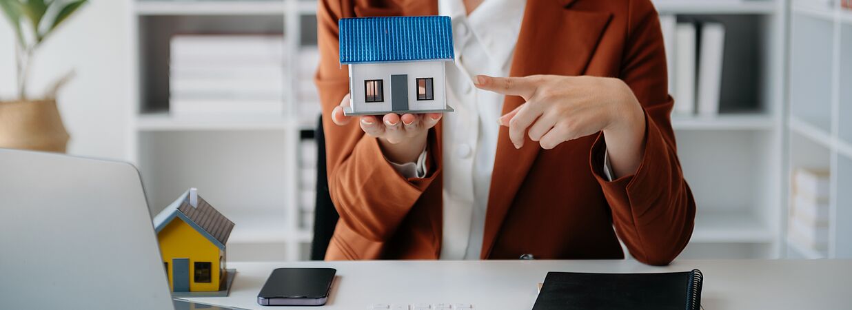 Frau zeigt auf kleines Haus, Rechner und Laptop