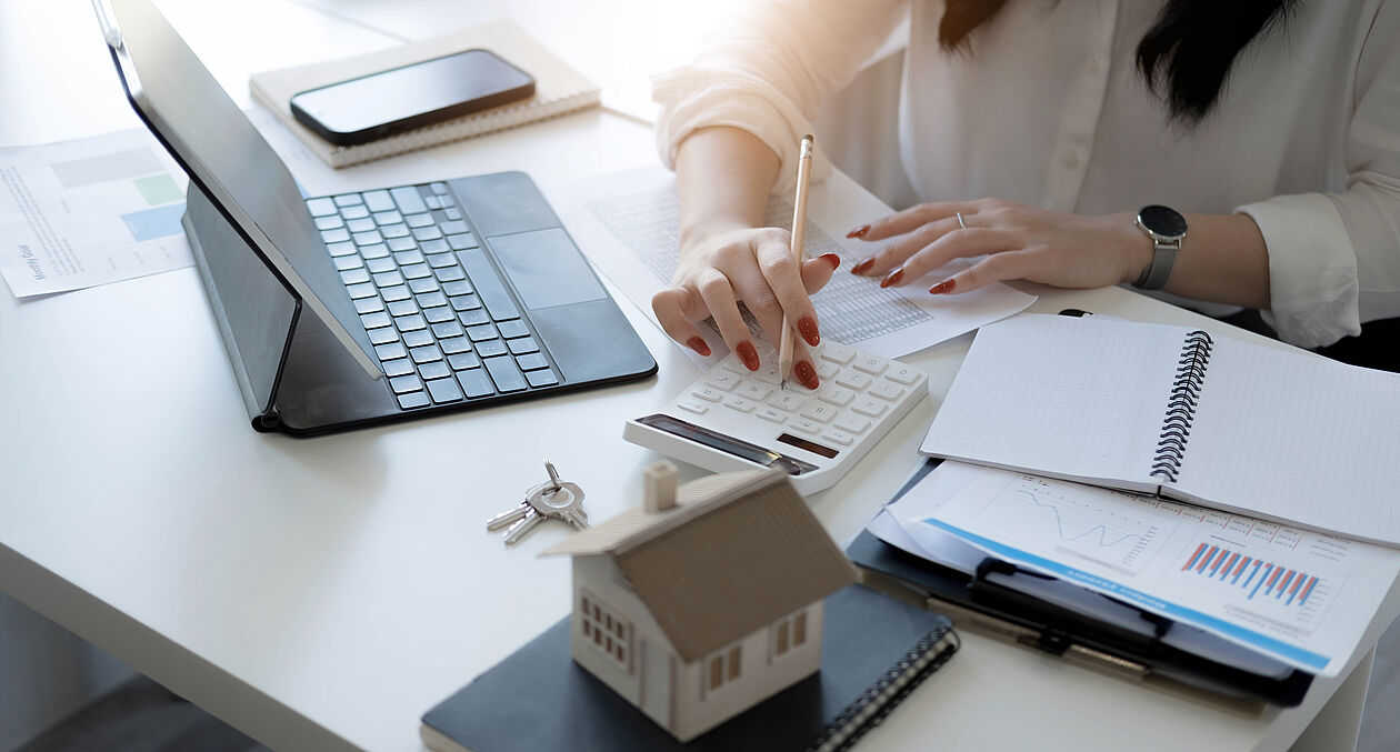 Eine Person berechnet den Immobilienwert mit Taschenrechner, Unterlagen und Hausmodell.