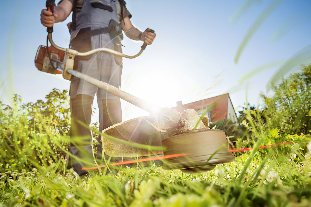 Person mäht hohen Rasen mit Motorsense in einem sonnigen Gartengrundstück.