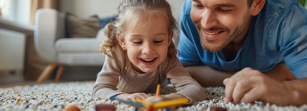 Vater und Tochter spielen lachend im Wohnzimmer und genießen ein sicheres Zuhause.