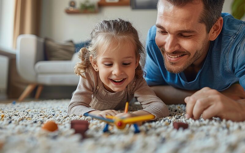 Vater und Tochter spielen lachend im Wohnzimmer und genießen ein sicheres Zuhause.