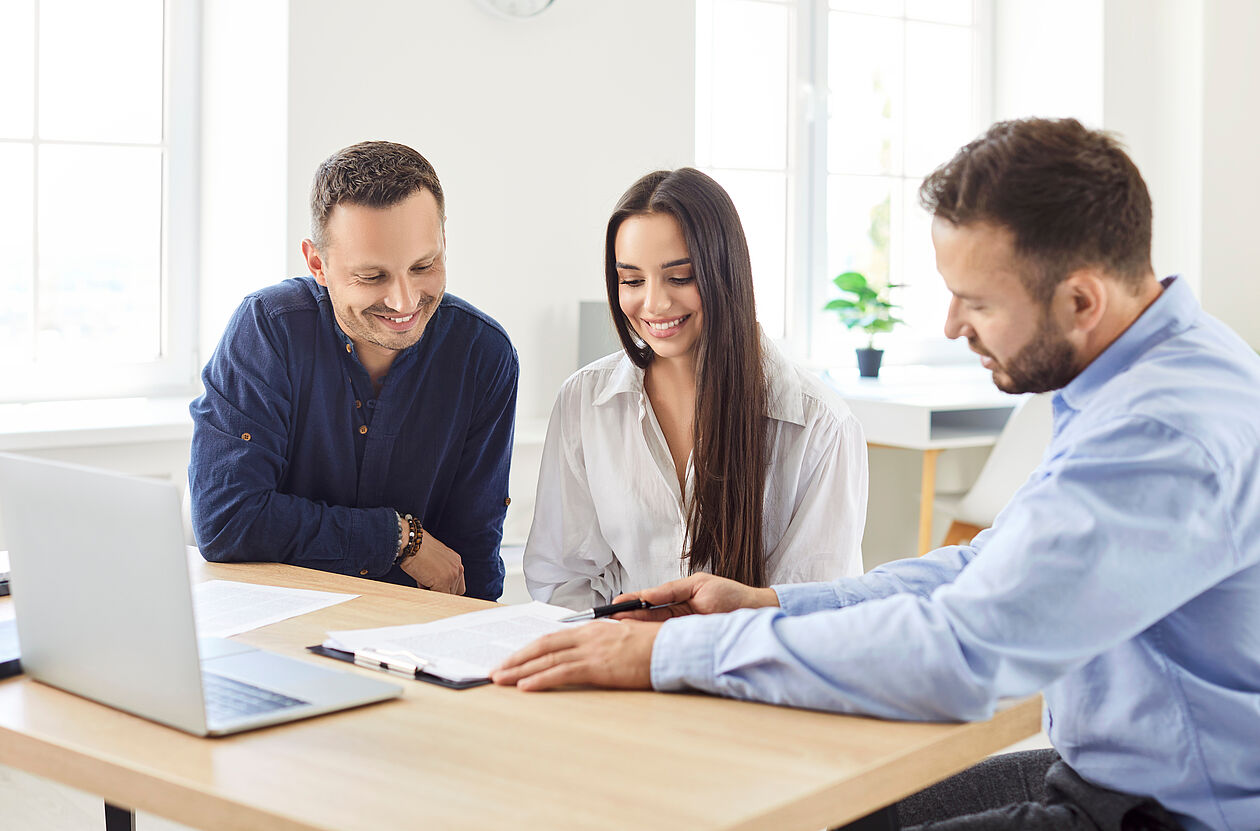 Junges Paar sitzt mit einem Immobilienberater am Tisch und prüft gemeinsam Vertragsunterlagen.