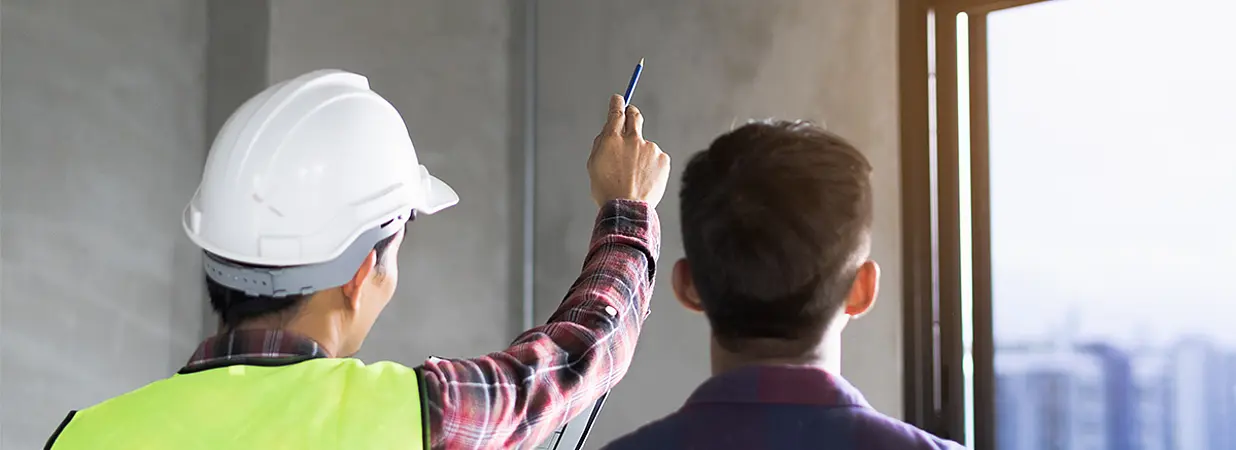 Ein Bauleiter zeigt auf eine Wand, während ein Kunde zuhört, sinnbildlich für Beratung und Planung bei der Umbaufinanzierung.