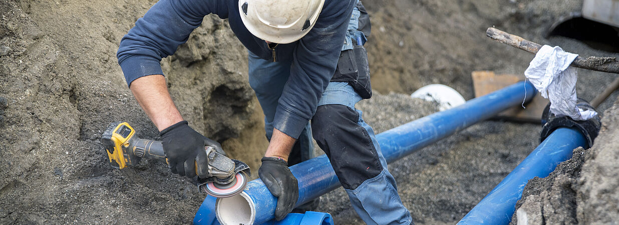 Ein Bauarbeiter mit Schutzhelm arbeitet in einer Baugrube an blauen Wasserrohren mit einem Winkelschleifer.