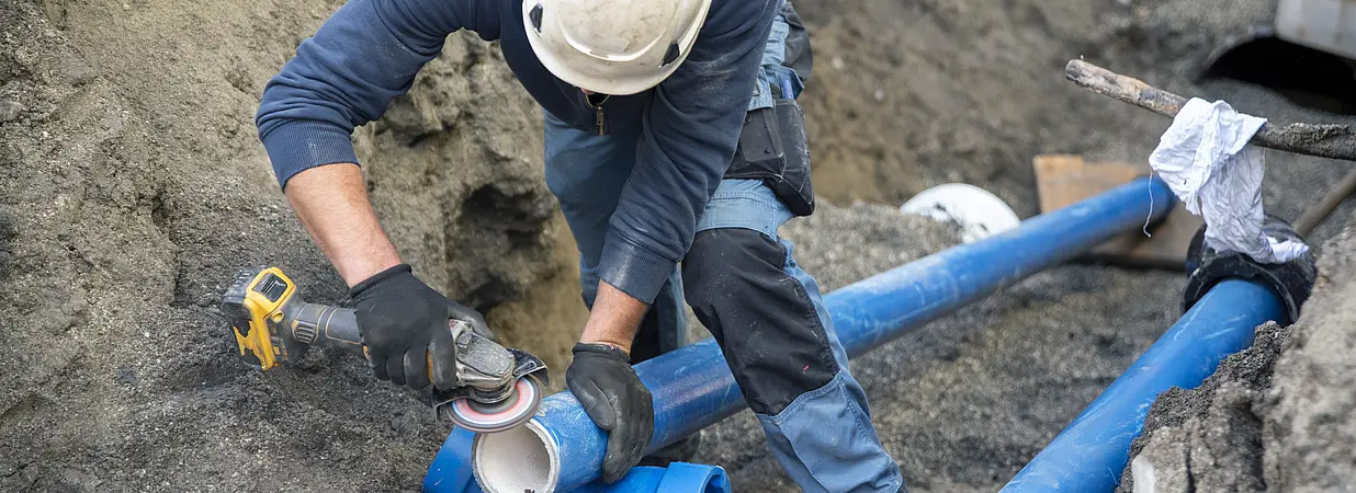 Ein Bauarbeiter mit Schutzhelm arbeitet in einer Baugrube an blauen Wasserrohren mit einem Winkelschleifer.