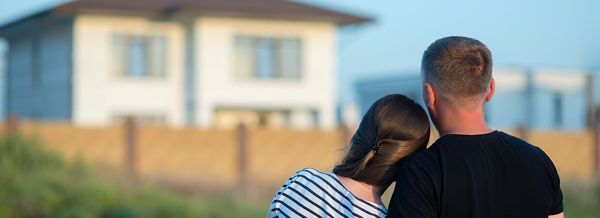 Ein Paar umarmt sich und blickt auf ein Einfamilienhaus in der Ferne.