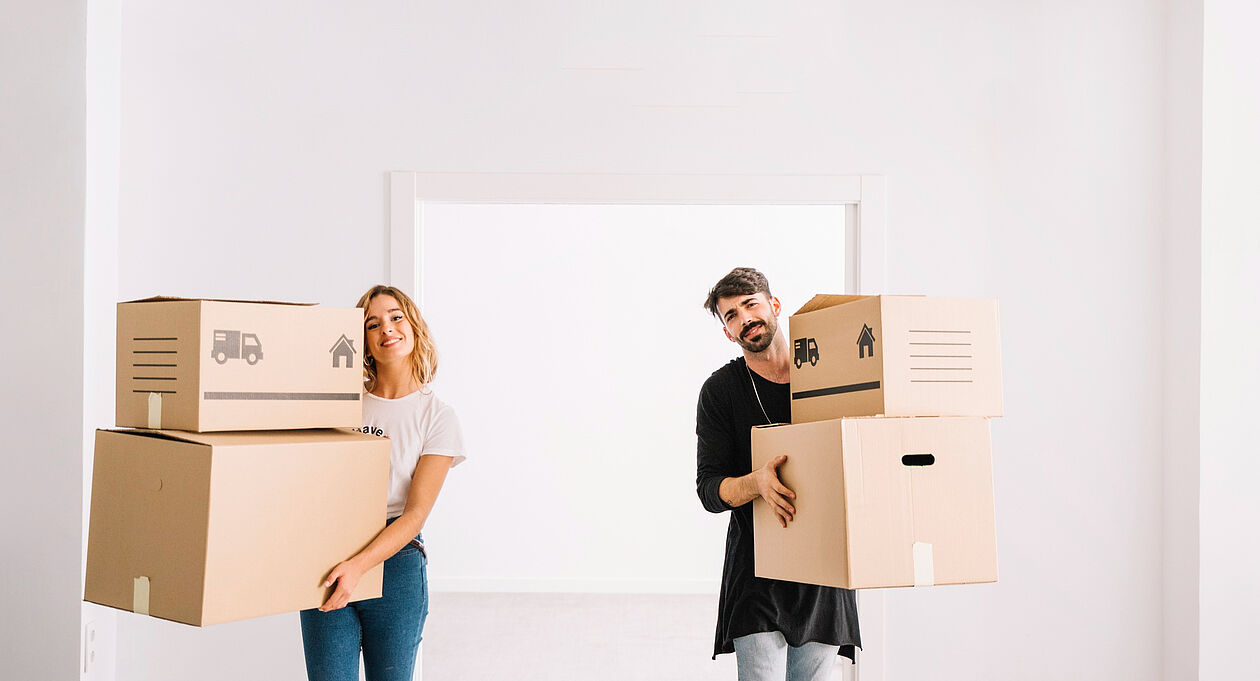 Ein junges Paar trägt lachend mehrere Umzugskartons in eine helle, leere Wohnung. Die Szene vermittelt Freude und Aufbruchsstimmung beim Einzug in ein neues Zuhause.