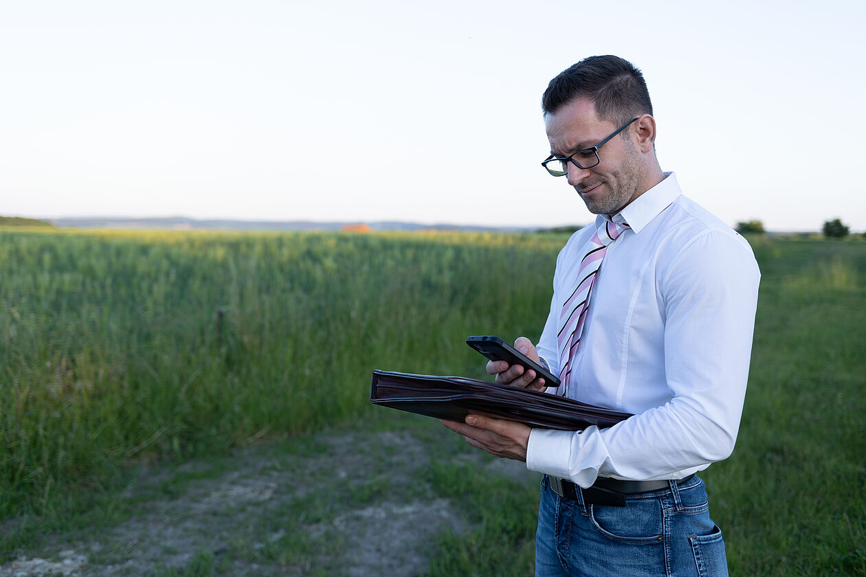Mann im weißen Hemd prüft mit Smartphone und Mappe ein Baugrundstück auf dem Land.