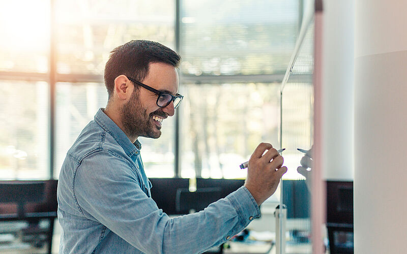 Mann mit Brille an einem White Board schreibt etwas auf.