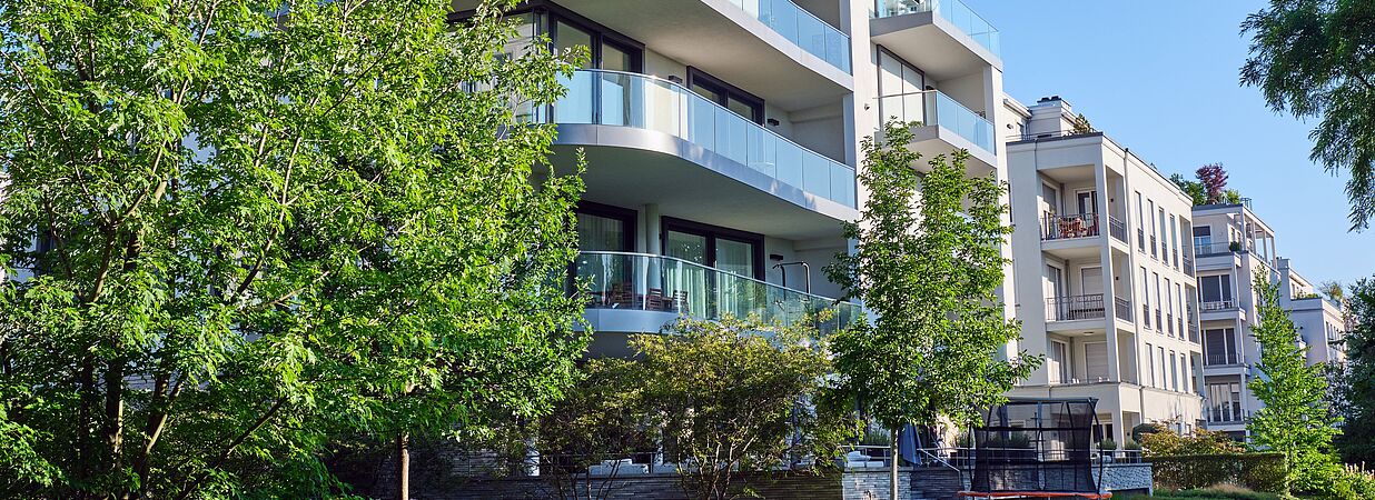Moderne, mehrstöckige Neubauwohnungen mit Glasbalkonen in grüner, urbaner Wohngegend bei Sonnenschein.