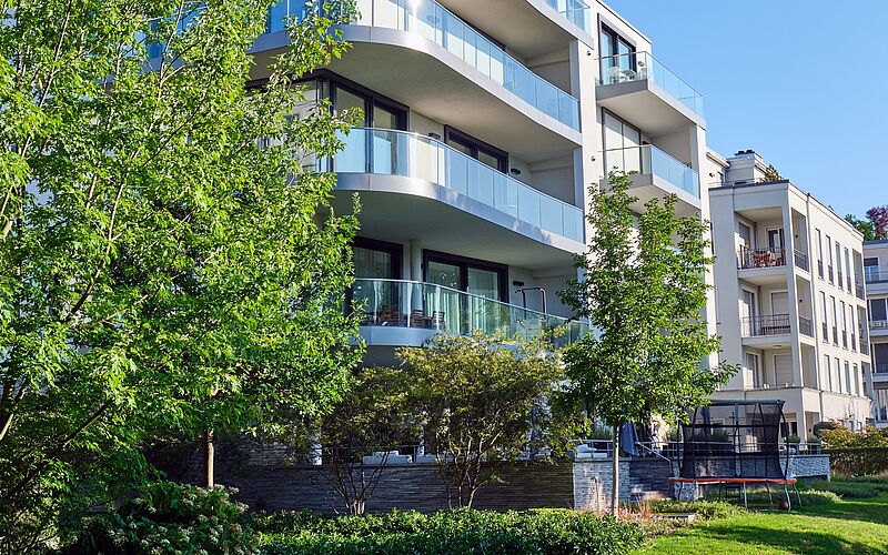 Moderne, mehrstöckige Neubauwohnungen mit Glasbalkonen in grüner, urbaner Wohngegend bei Sonnenschein.