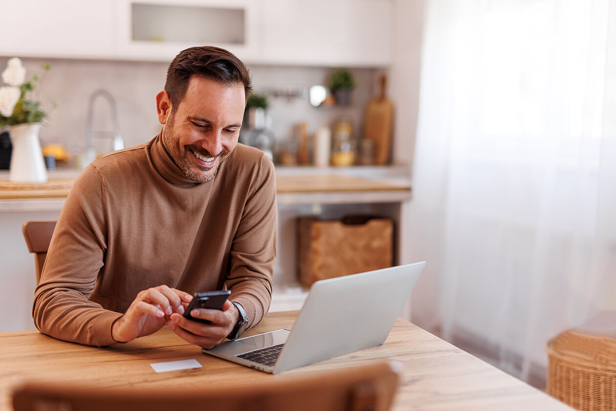 Mann sitzt entspannt in moderner Wohnung am Tisch mit Laptop und Smartphone.