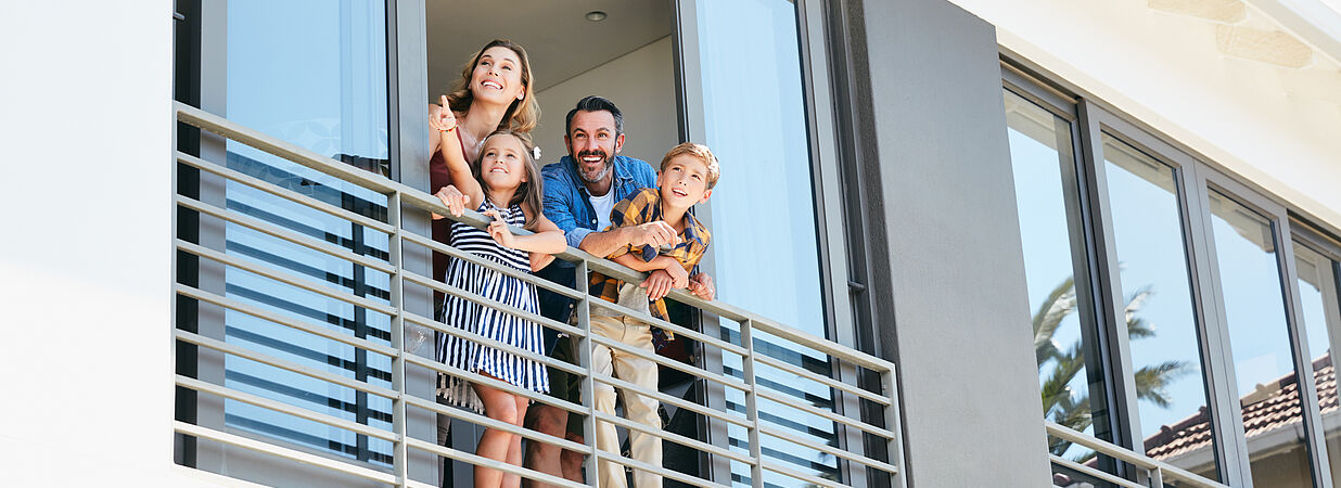 Junge Familie steht lachend auf dem Balkon einer modernen Wohnung und blickt nach draußen.