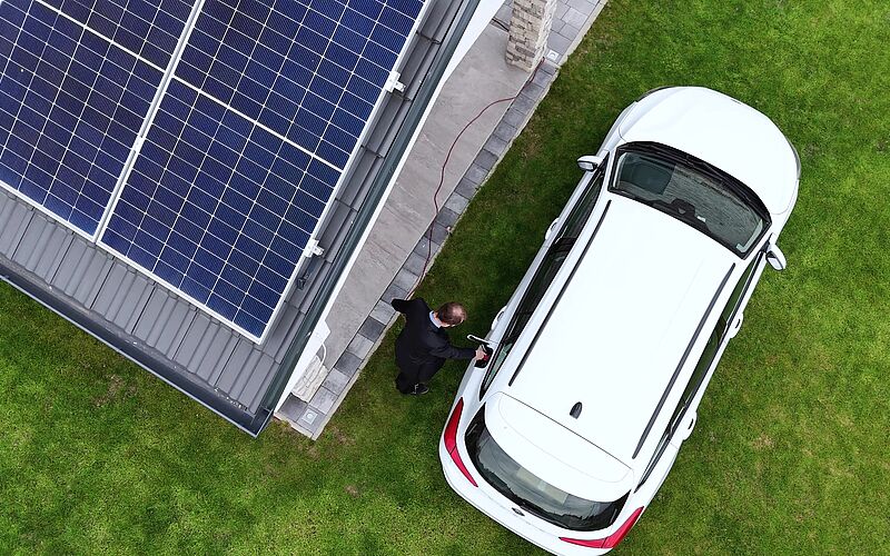 Drohnenansicht eines Hauses mit Photovoltaikanlage und weißem Elektroauto.