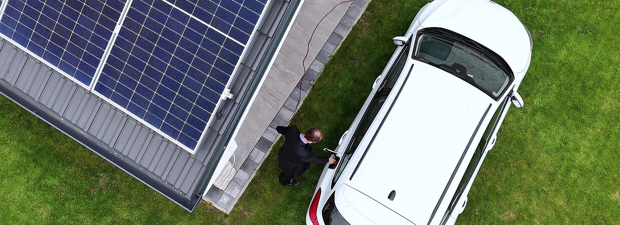 Drohnenansicht eines Hauses mit Photovoltaikanlage und weißem Elektroauto.