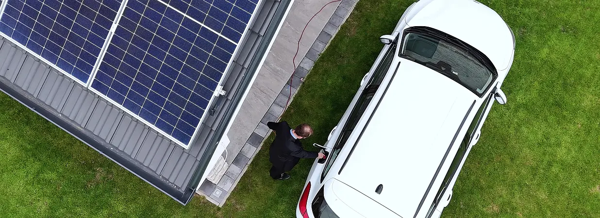 Drohnenansicht eines Hauses mit Photovoltaikanlage und weißem Elektroauto.