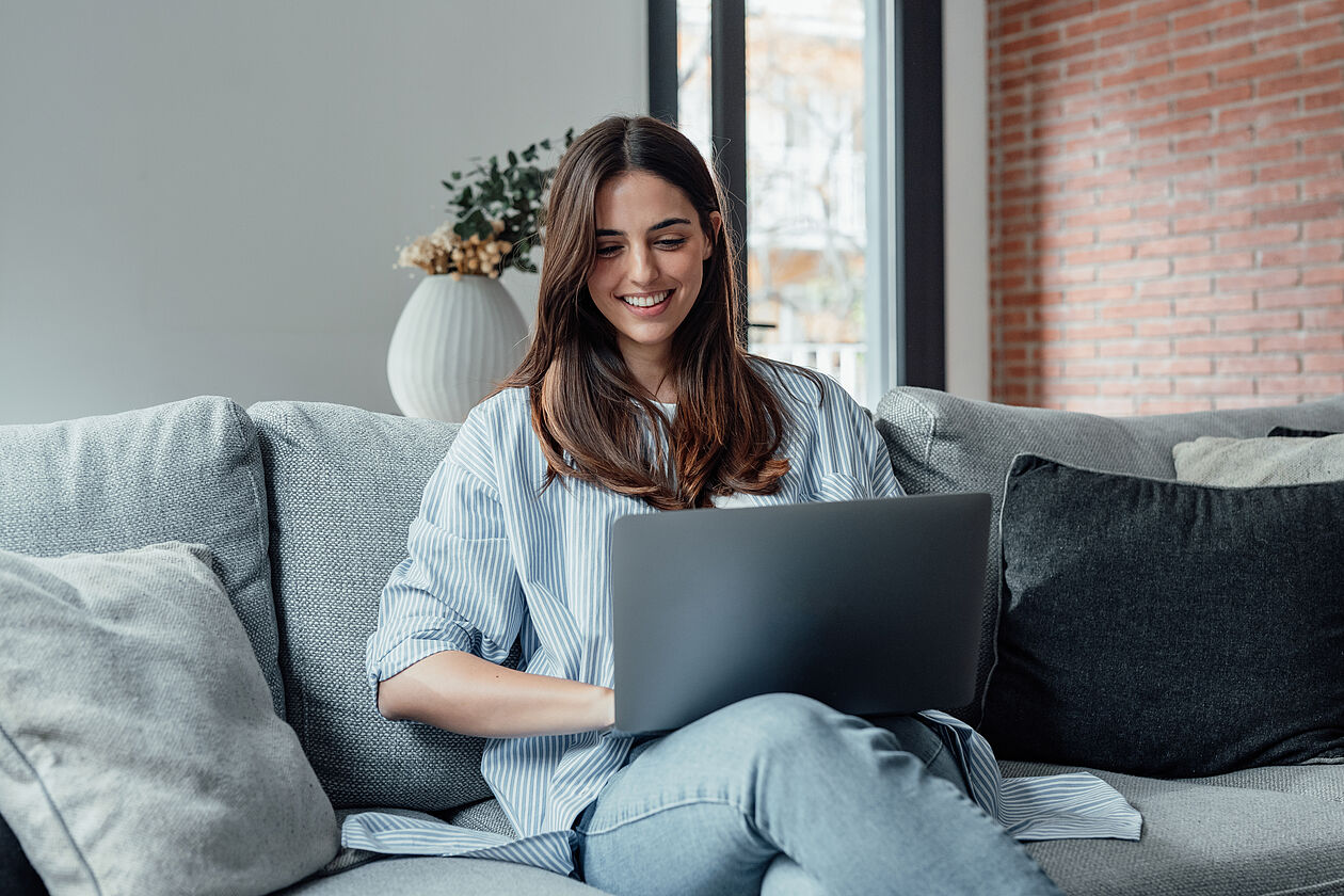 Junge Frau sitzt entspannt auf dem Sofa und nutzt Laptop zur Finanzplanung.