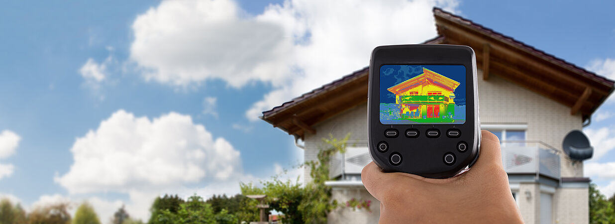 Hand hält Wärmebildkamera vor ein Einfamilienhaus, auf dem Display ist ein Thermogramm mit farblich sichtbaren Wärmeverlusten zu sehen.
