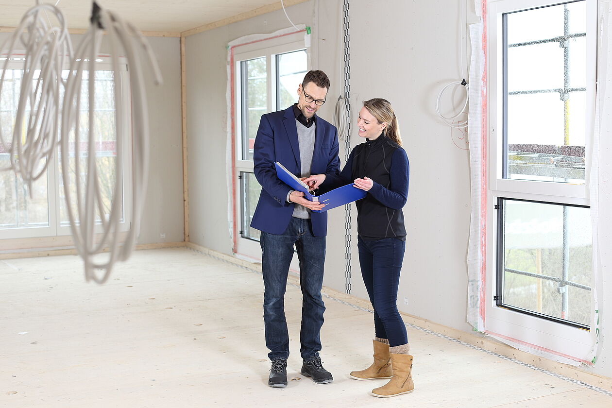 Sachverständiger und Kundin begutachten ein Neubauobjekt mit Bauunterlagen auf der Baustelle.