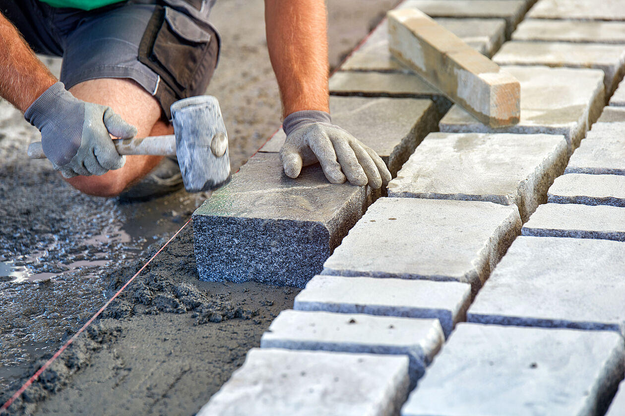 Ein Arbeiter mit Handschuhen klopft mit einem Hammer auf einen Terrassenstein um ihn im nassen Beton darunter zu verankern.
