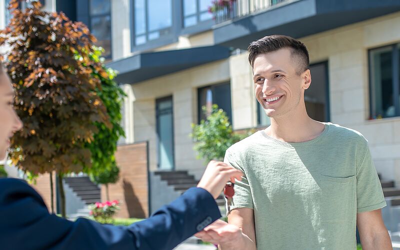 Lächelnder Mann erhält vor einem modernen Wohngebäude die Schlüssel zu seiner neuen Immobilie.